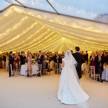 Bride and groom entering a marquee with a fairy light canopy covering the dining section, and another section with clear roofs